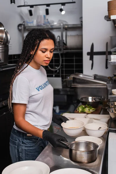 African american woman in t-shirt with volunteer lettering holding saucepan near plastic bowls — Foto stock