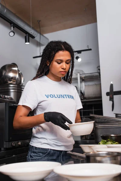 African american woman in t-shirt with volunteer lettering holding plastic bowl with soup in kitchen — Stockfoto