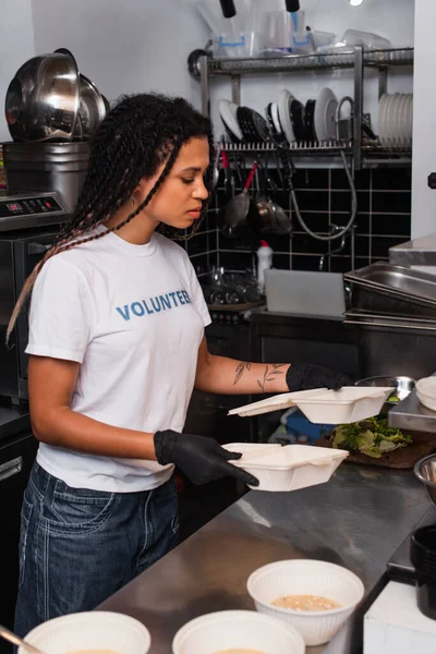 Tattooed african american woman in t-shirt with volunteer lettering holding plastic containers in kitchen — стоковое фото