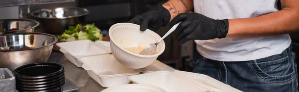 Partial view of tattooed african american volunteer holding plastic bowl with soup in kitchen, banner — стоковое фото