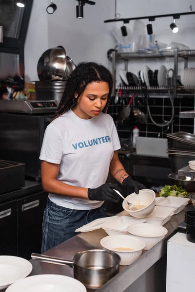 Tattooed african american volunteer in t-shirt with lettering holding plastic container with soup and fork in kitchen — Photo de stock