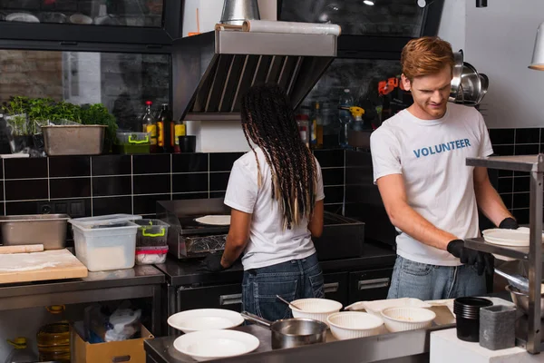 Happy redhead volunteer cooking near african american woman in kitchen — Foto stock