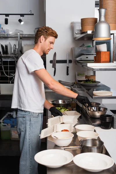 Redhead volunteer in latex gloves cooking near plastic containers in kitchen — Fotografia de Stock