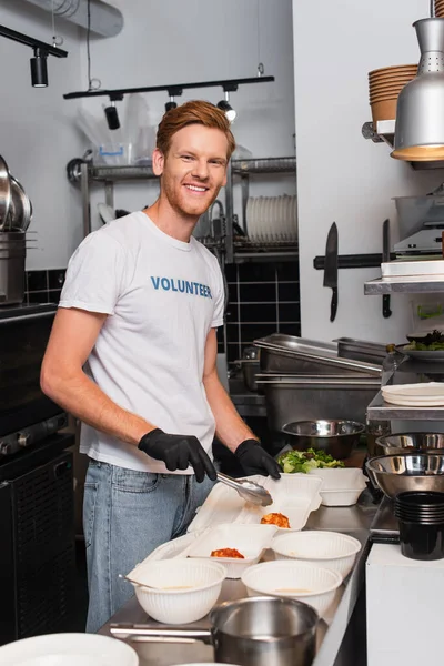 Happy and redhead volunteer in latex gloves holding tweezers near plastic containers in kitchen — Foto stock