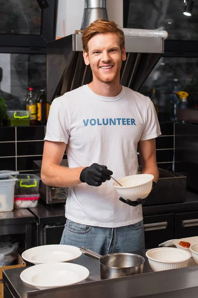 Happy and redhead volunteer in latex gloves holding plastic bowl in kitchen — Fotografia de Stock
