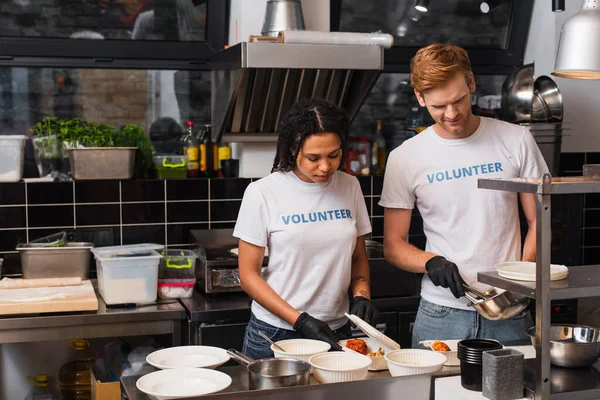 Multiethnic social workers in latex gloves putting prepared food into plastic containers — Stock Photo