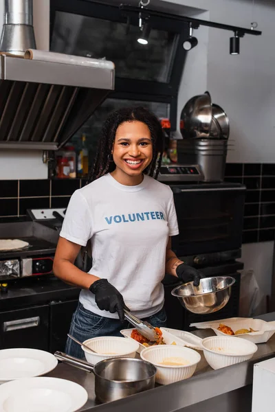 Happy african american social worker putting prepared meal into plastic containers in kitchen — Photo de stock