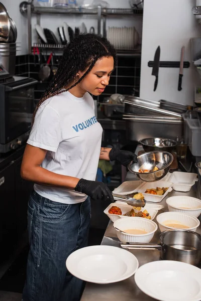 Young african american social worker putting prepared meal into plastic containers in kitchen — Stock Photo