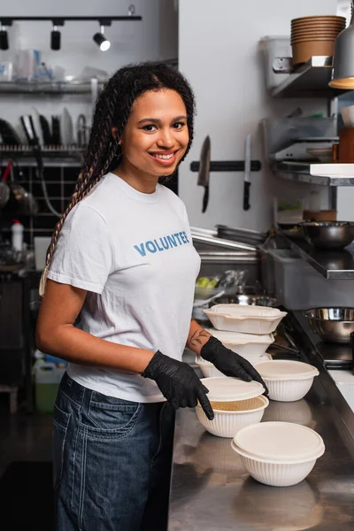 Cheerful african american social worker covering plastic container with cup in kitchen - foto de stock