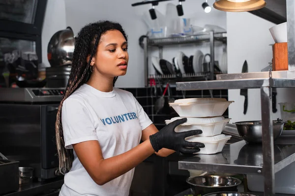 Young african american social worker holding plastic containers in kitchen - foto de stock