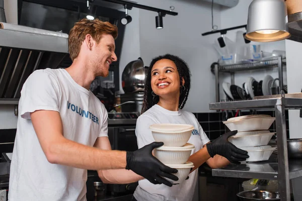 Smiling multiethnic volunteers in t-shirts holding plastic containers with food in kitchen — стоковое фото