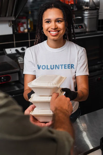 Happy african american volunteer giving plastic containers with food to man - foto de stock