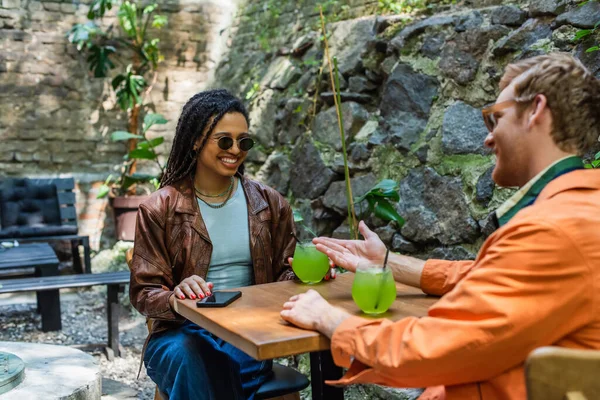 Cheerful man pointing at happy african american friend in sunglasses near cocktail — Photo de stock