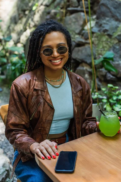 Happy african american woman in sunglasses holding glass with green alcohol cocktail in outdoor terrace — Stockfoto