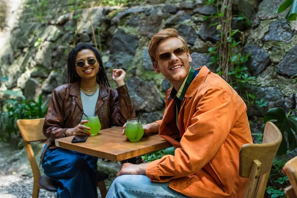 Happy multiethnic friends in sunglasses and jackets holding glasses with green alcohol cocktail in outdoor terrace — Fotografia de Stock