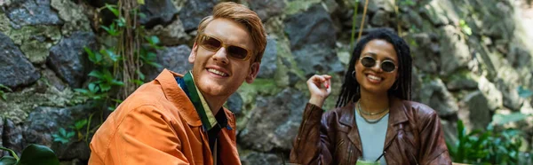 Happy multiethnic friends in sunglasses and jackets smiling in outdoor terrace, banner — стоковое фото