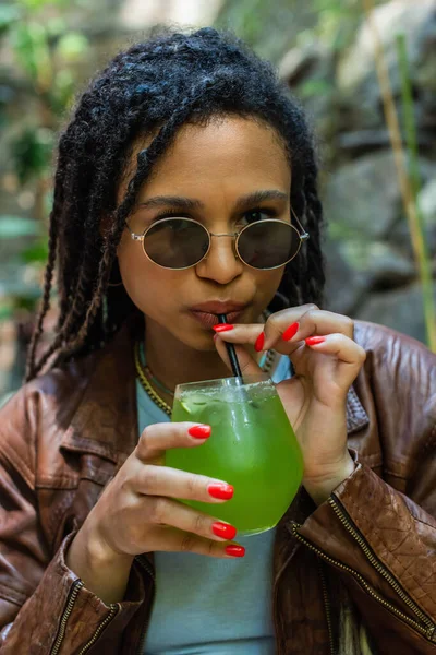 African american woman in stylish sunglasses holding glass and drinking green alcohol cocktail through paper straw — Fotografia de Stock