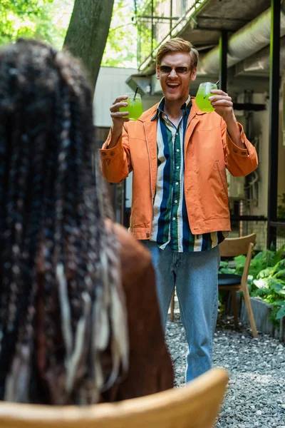 Cheerful redhead man in sunglasses holding glasses with green alcohol cocktail near blurred friend - foto de stock