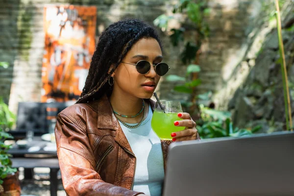 Молодий афроамериканський фрілансер в сонцезахисних окулярах використовує ноутбук і тримає скло з коктейлем на відкритій терасі — стокове фото