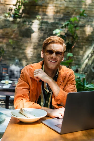 Happy redhead freelancer in sunglasses using laptop near prepared lunch on plate — Photo de stock