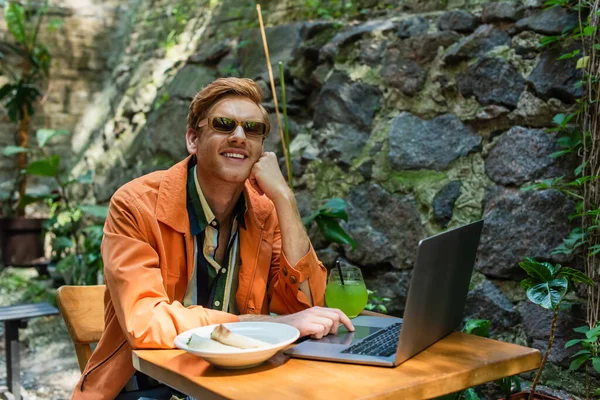 Happy redhead freelancer in sunglasses using laptop near prepared lunch and cocktail on table — Photo de stock