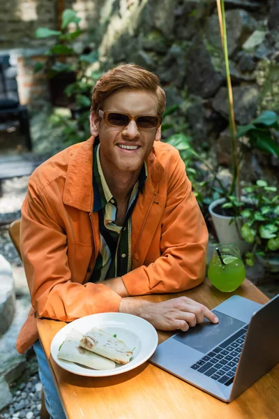 Cheerful redhead freelancer in sunglasses using laptop near prepared lunch and cocktail on table — Stock Photo