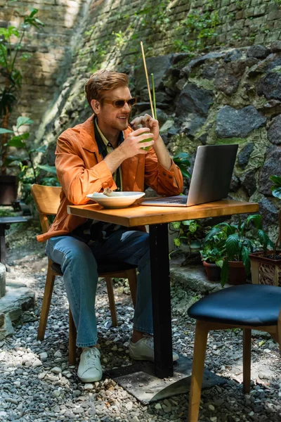 Cheerful redhead freelancer in sunglasses holding glass with cocktail near laptop and quesadillas in outdoor terrace — Stockfoto