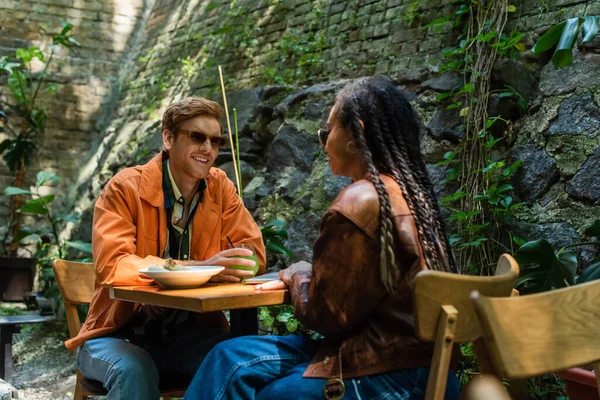 Cheerful redhead man holding glass with lemonade near prepared lunch and african american friend in cafe terrace - foto de stock