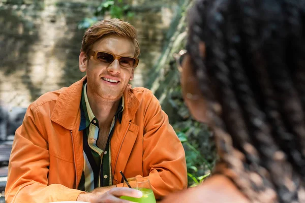 Cheerful redhead man holding glass with lemonade near blurred african american friend in outdoor terrace — Stockfoto