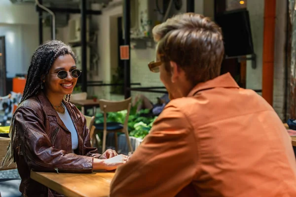 Щаслива афроамериканка в сонцезахисних окулярах дивиться на розмитого друга на терасі відкритого кафе — стокове фото