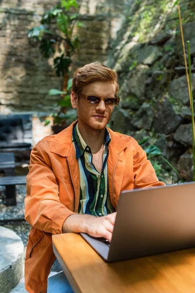 Redhead man in sunglasses working remotely on laptop while sitting in outdoor terrace - foto de stock