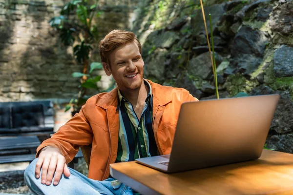 Happy redhead man looking at laptop while sitting in outdoor terrace — стоковое фото