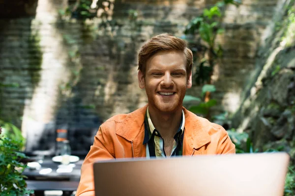Joyful redhead man looking at laptop while working remotely in outdoor terrace - foto de stock