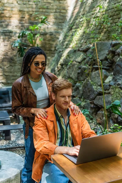 Happy african american woman touching shoulders of redhead man using laptop in outdoor terrace — Stock Photo