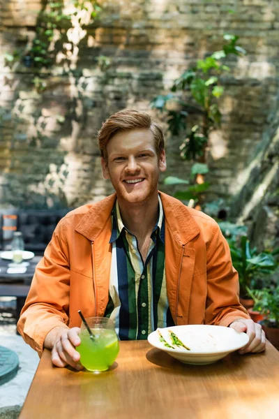 Happy redhead man near prepared lunch and glass with cocktail in outdoor cafe terrace — стоковое фото