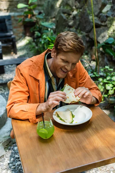Happy redhead man eating quesadilla near glass with cocktail in outdoor terrace — стоковое фото