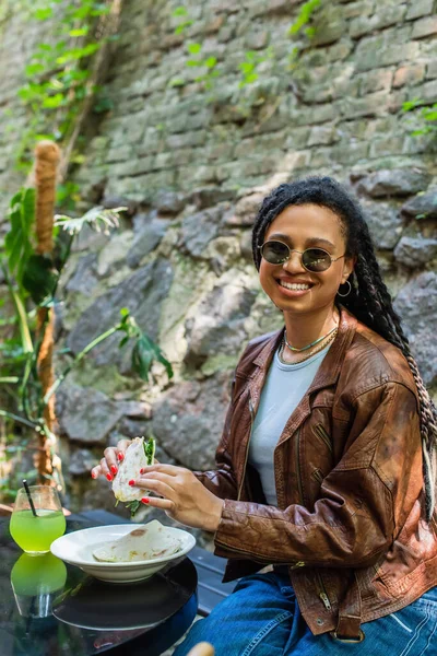 Cheerful african american woman holding quesadilla near glass with cocktail in outdoor cafe terrace — Photo de stock