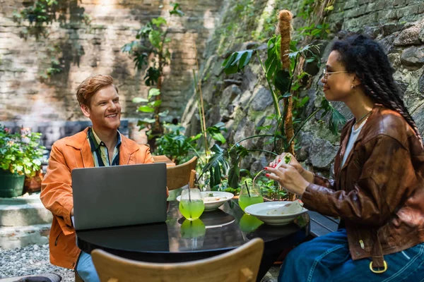 African american woman holding quesadilla near happy freelancer friend using laptop in outdoor terrace — Fotografia de Stock
