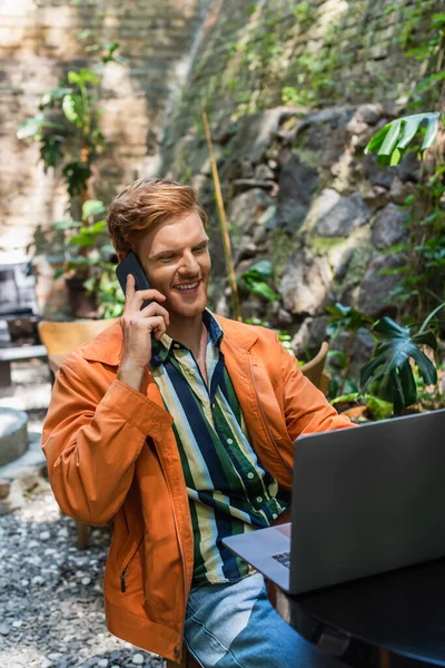 Cheerful redhead man talking on smartphone while using laptop in cafe terrace — стоковое фото