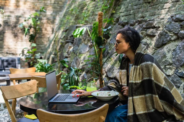 African american freelancer covered in blanket using laptop and having lunch in cafe on terrace — Fotografia de Stock
