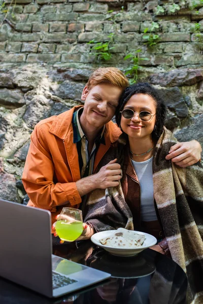 Happy redhead man hugging african american friend covered in blanket while having lunch in cafe near laptop — Stockfoto