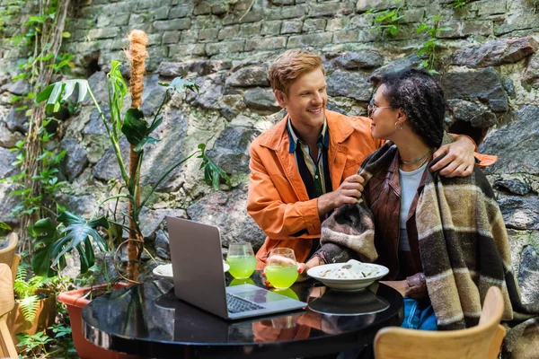 Happy redhead man looking at african american woman covered in blanket while having lunch in cafe near laptop — стоковое фото