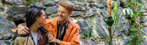 Happy redhead man looking at african american woman covered in blanket in cafe on terrace, banner - foto de stock