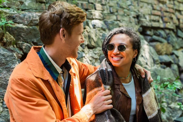 Redhead man hugging happy african american woman covered in blanket in cafe on terrace — стоковое фото