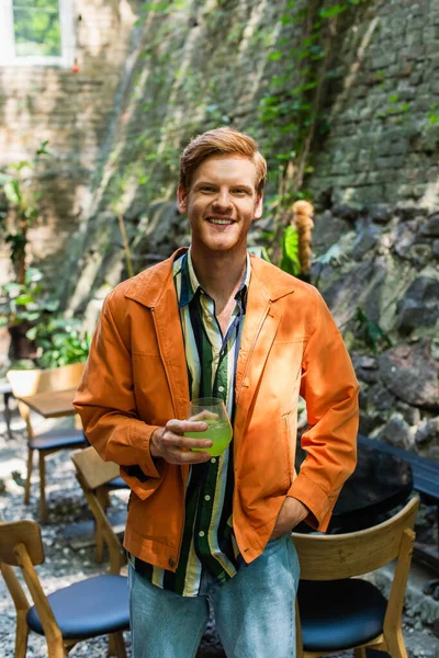 Cheerful redhead man in orange jacket standing with hand in pocket and holding glass with cocktail — Stock Photo