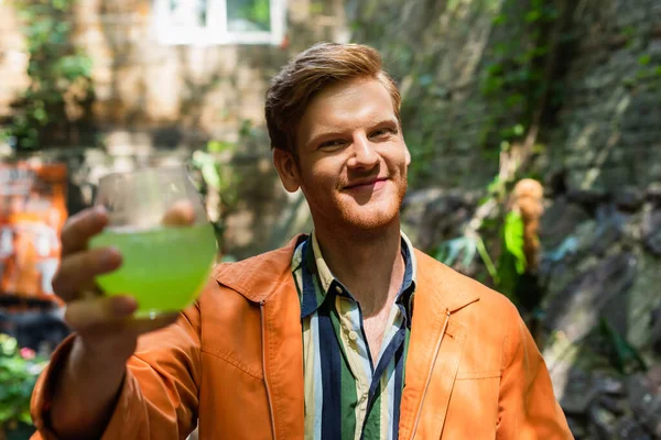 Cheerful redhead man in orange jacket holding blurred glass with cocktail — стоковое фото