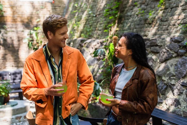 Happy redhead man and african american woman holding glasses with green cocktail outside — Stock Photo