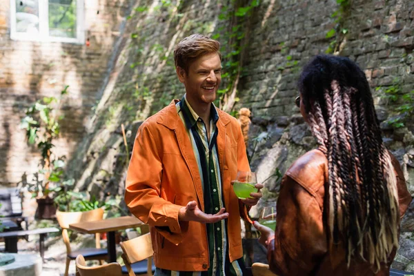 Happy redhead man looking at african american woman while gesturing and holding glass with green cocktail outside - foto de stock