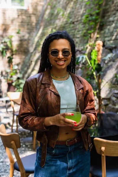 Happy african american woman in sunglasses holding glass with green cocktail outside — Photo de stock