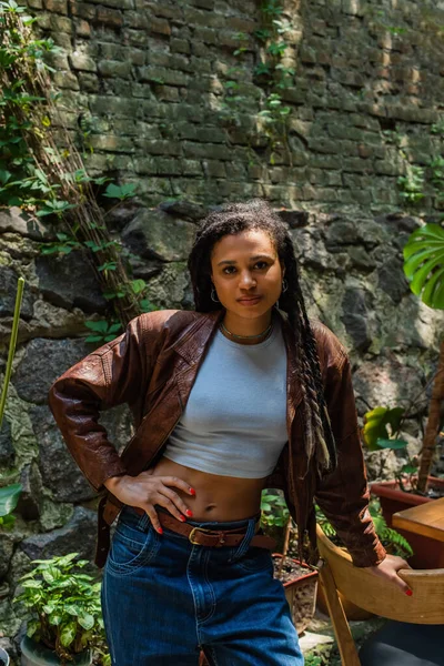 Young african american woman in brown leather jacket standing with hand on hip - foto de stock